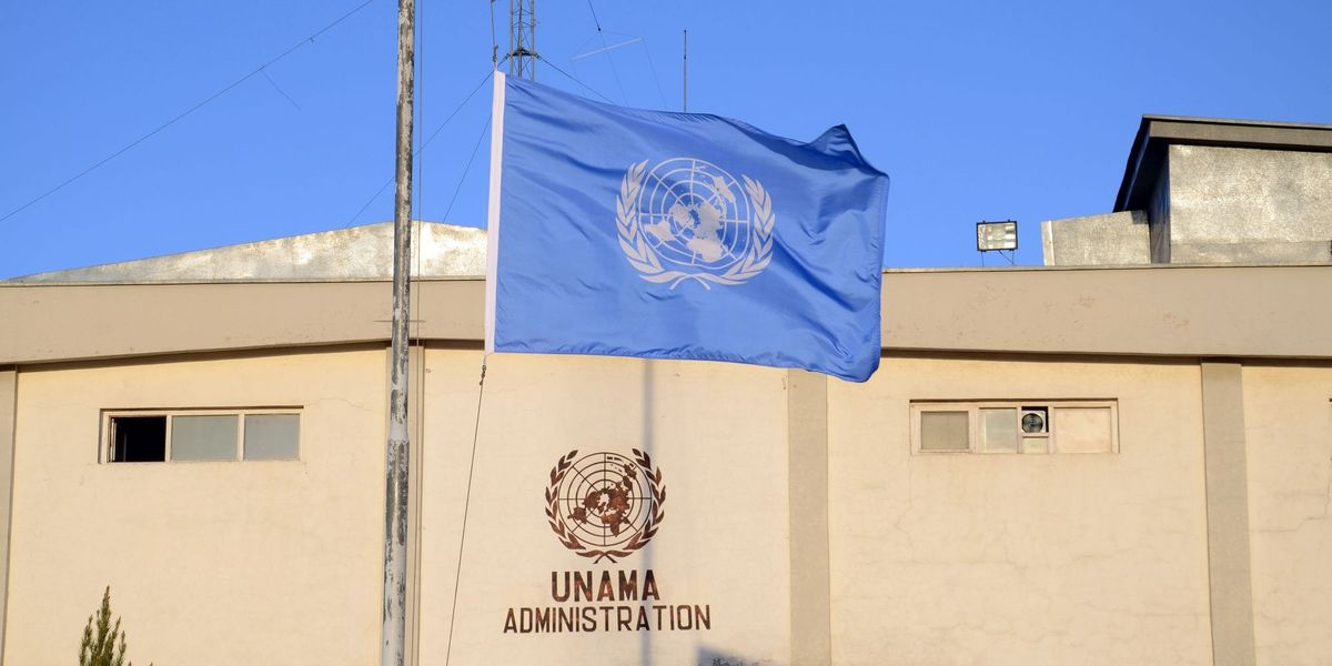 Vor einem sandfarbenen Gebäude mit dem UNAMA-Logo weht die blaue UN-Flagee auf Halbmast.
