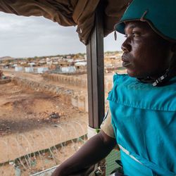 Ein Blauhelmsoldat der UNAMID-Mission wacht über ein Camp von Geflüchteten in Darfur, 2014 in einem Wachturm sitzt ein Mann mit blauem UN-Helm und UN-Weste und blickt auf eine Wüstenlandschaft mit Hütten.