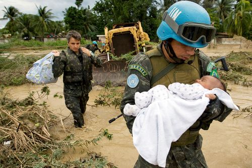 Ein Soldat mit blauem Helm trägt ein Baby durch ein überschwemmtes Gebiet.