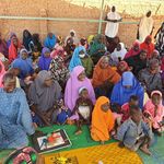 Eine Gruppen von Frauen und Kinder sowie ein Mann sitzen auf dem Boden, vielen lachen und schauen nach rechts.