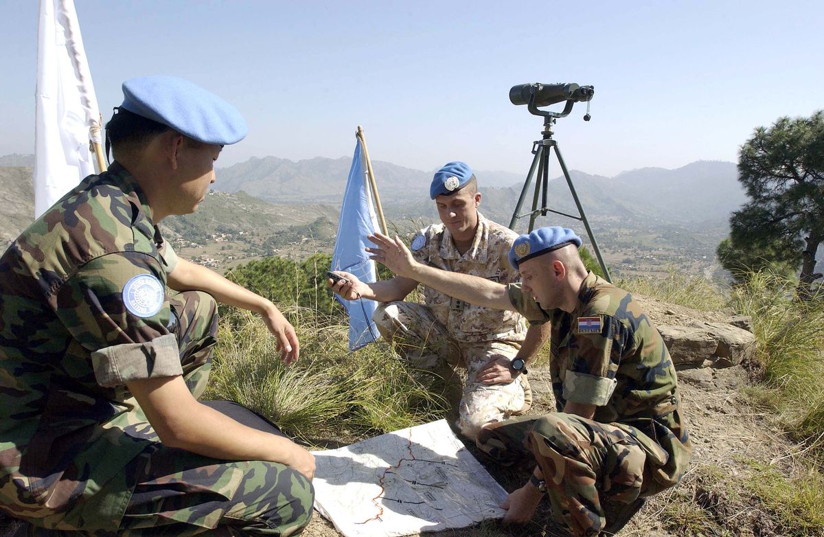 Drei Soldaten sitzen auf einem Plateau. Zwischen ihnen liegt eine Landkarte, hinter ihnen sind zwei Flaggen, eine der UN. Im Hintergrund Gebirge und Täler.
