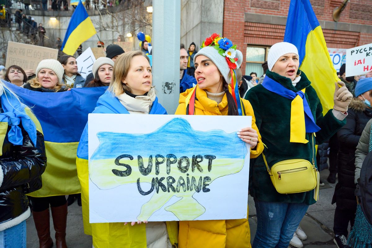 Demonstrantinnen halten ein Schild mit der Aufschrift "Support Ukraine", im Hintergrund sieht man die ukrainische Flagge.